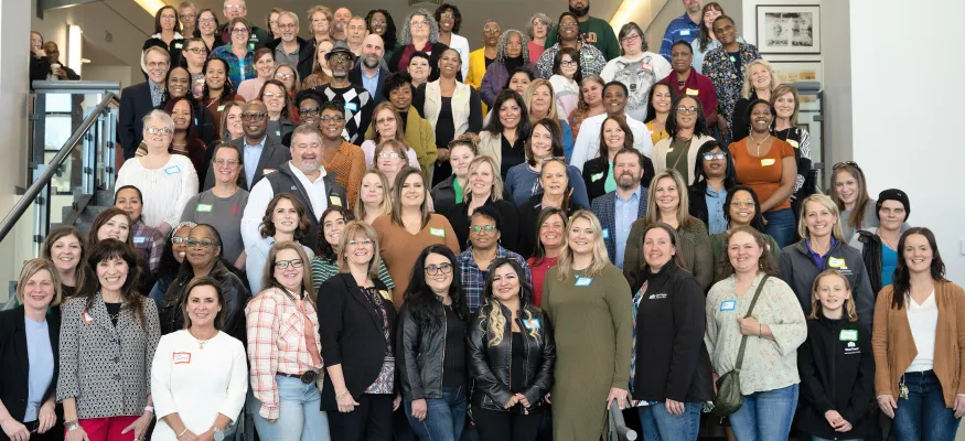 A large, diverse group of people stand and sit on a staircase inside a building, posing for a group photo. Some individuals wear nametags and are dressed casually.