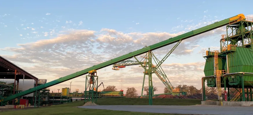 A green, industrial conveyor belt system is shown extending across an outdoor facility, with metal structures and large containers underneath a partly cloudy sky.