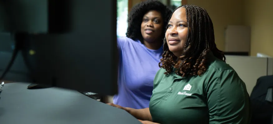 Two people are working at computers in an office. One is wearing a purple shirt and the other a green shirt. They appear focused on their tasks.