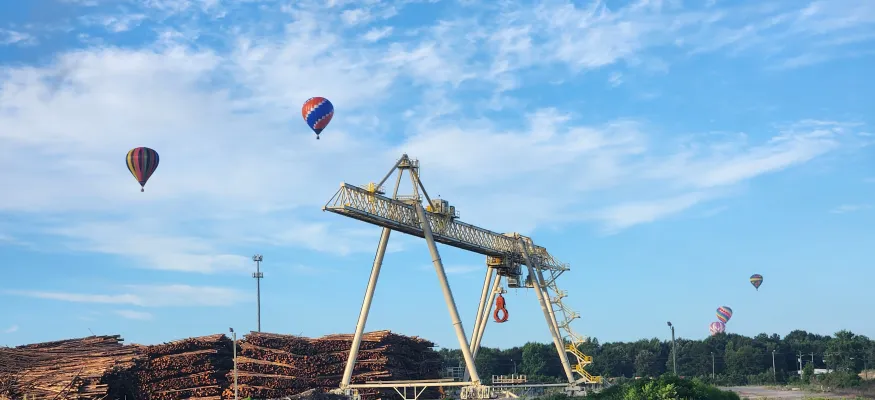 A large industrial crane is stationed near stacks of logs by a water body. Several hot air balloons are floating in the sky above, against a backdrop of trees and a clear blue sky.