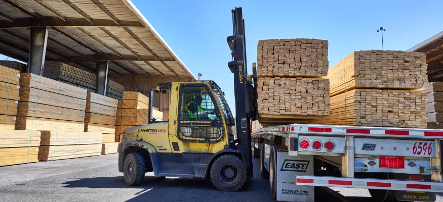 A forklift operator loads a large stack of lumber onto a flatbed trailer in an outdoor lumber yard, surrounded by stacks of wooden planks under a clear blue sky.