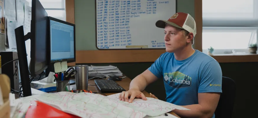 A West Fraser worker wearing a cap and a blue shirt sits at a desk with a computer, keyboard, and maps, surrounded by office materials and a whiteboard in the background.