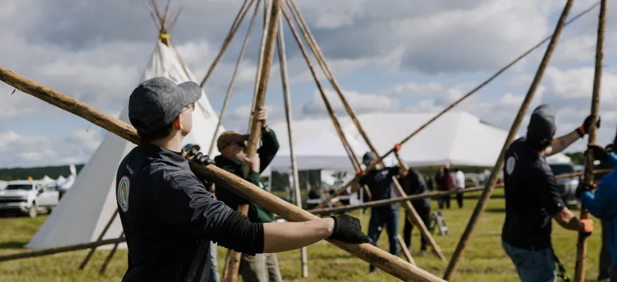 Several people work together to set up wooden poles for a large tent structure or teepee frame in an open field on a cloudy day.