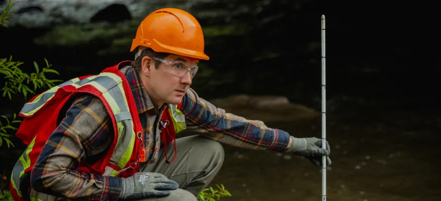 Person in a safety vest and helmet crouches by a stream, holding a measuring stick near a bridge.