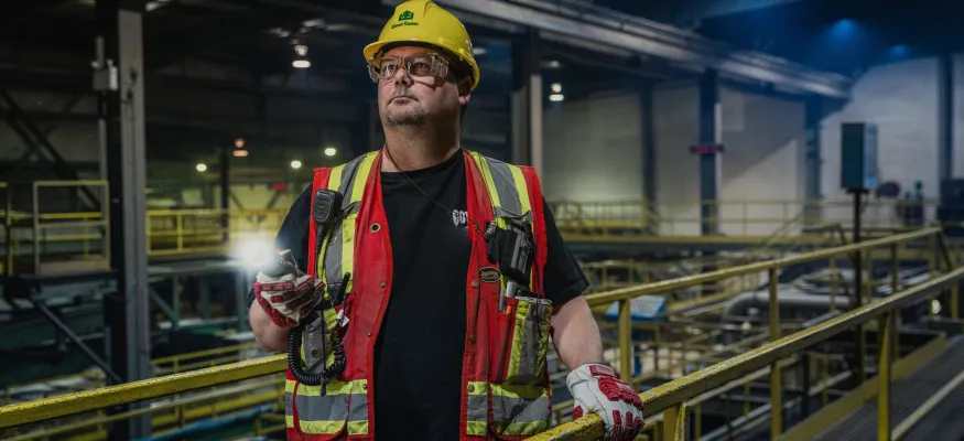 A worker in a hard hat and safety vest stands in an industrial facility, holding a flashlight. The background includes metal railings and machinery, indicating a factory or warehouse environment.