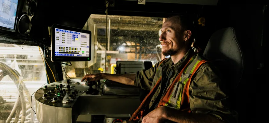 Person in a high-visibility vest operates machinery, sitting in a control room with multiple screens and control panels.