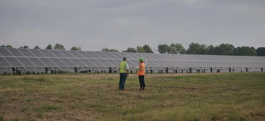 Two people in safety gear stand in a grassy field near large rows of solar panels under a cloudy sky.