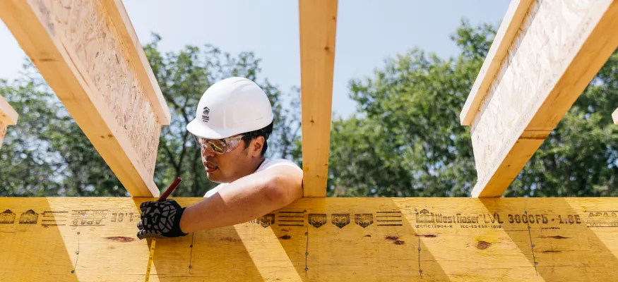 A construction worker wearing a white hard hat and safety glasses measures and marks a wooden beam at a construction site.