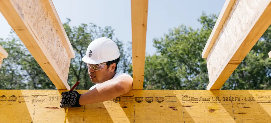 A construction worker wearing a white hard hat and safety glasses measures and marks a wooden beam at a construction site.