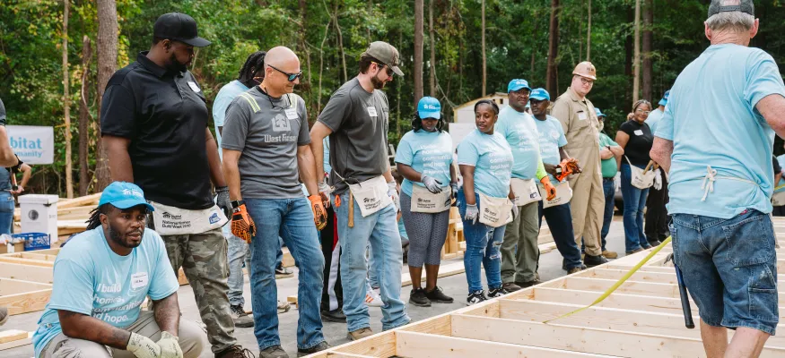 A group of people wearing casual work clothes and teal hats gather around wooden frames on the ground in an outdoor construction area.