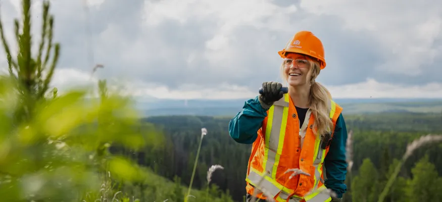 A person wearing an orange hard hat and safety vest stands in a green forested area, smiling and holding a radio.