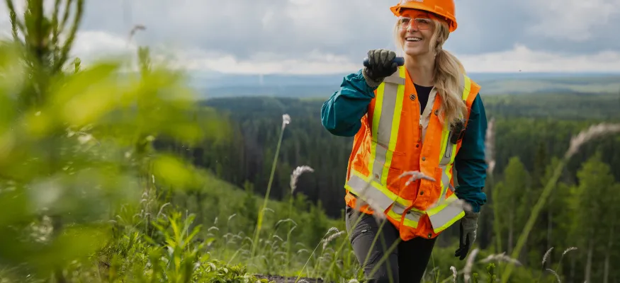 A person wearing an orange hard hat and safety vest stands in a green forested area, smiling and holding a radio.