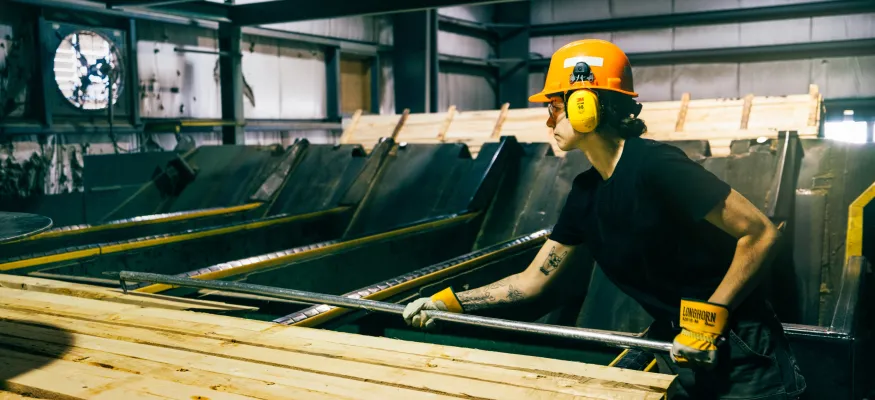 A worker wearing protective gear handles wooden planks in an industrial setting, with machinery visible in the background.