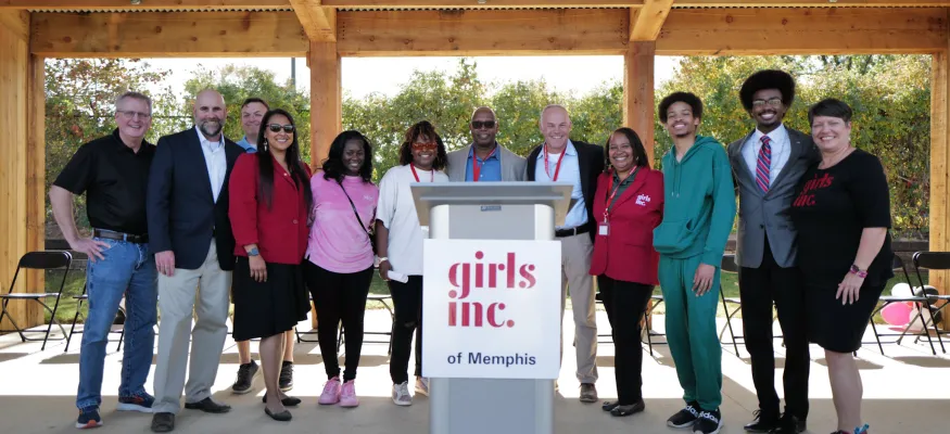 A group of people stands together under a wooden pavilion, with a podium labeled "Girls Inc. of Memphis" in front.