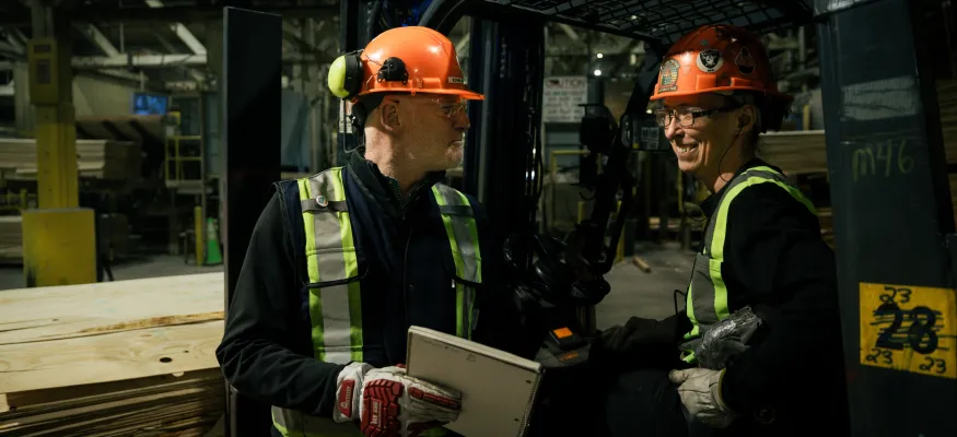 Two workers wearing hard hats and safety vests converse near a forklift in a lumber warehouse. One holds a tablet, and the other is seated on the forklift.
