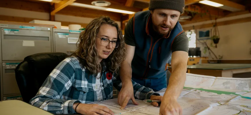 Two people reviewing large maps on a table. The woman is seated, wearing glasses, and the man is pointing at the map, wearing a beanie. They are in a room with wooden beams and filing cabinets.