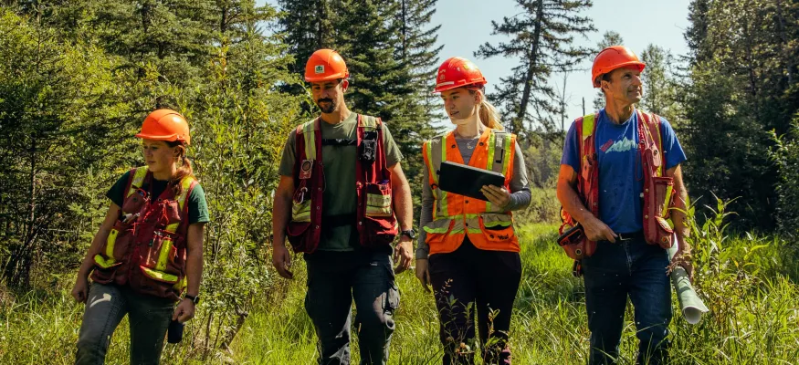 Four people wearing safety gear and helmets walk through a grassy area with tall trees in the background.