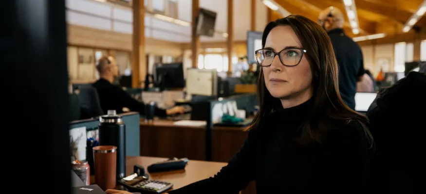 A woman with glasses works at a computer in an office with wooden ceilings. Other people are seen working in the background.