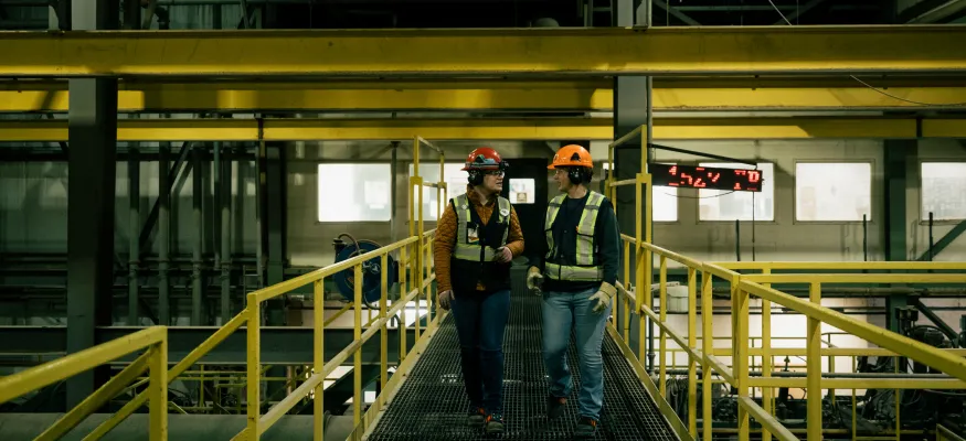 Two workers in safety gear walk on a metal platform inside an industrial facility with yellow railings and overhead structures.