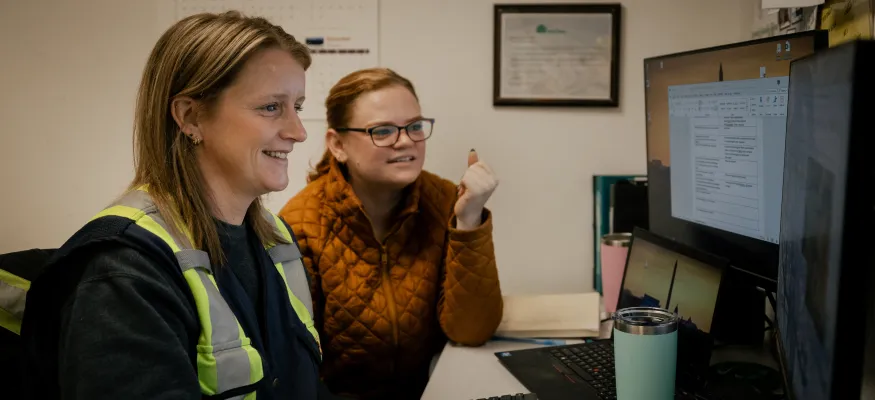 Two women sitting at a desk, looking at a computer screen. One wears a reflective vest. The wall behind them has a calendar and framed certificates.