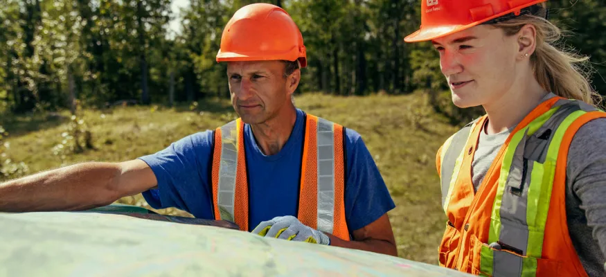 Two people wearing orange safety vests and hard hats look at a large map outdoors in a wooded area on a sunny day.
