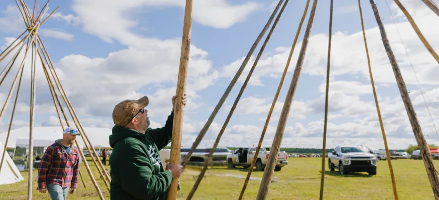 Two men assemble wooden poles in a grassy field under a partly cloudy sky, with trucks and tents in the background.