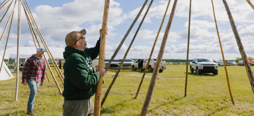 Two men assemble wooden poles in a grassy field under a partly cloudy sky, with trucks and tents in the background.