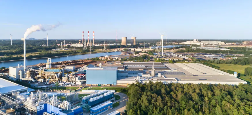 Aerial view of an industrial complex with factories, smokestacks emitting smoke, warehouses, wind turbines, and surrounding greenery under a clear sky.