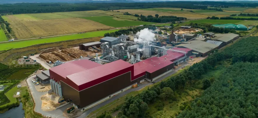 Aerial view of an industrial facility with red roofs emitting steam, surrounded by trees, fields, and stacks of logs in a rural landscape.