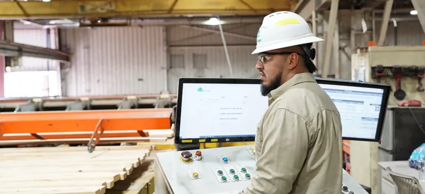 A worker wearing a hard hat and gloves operates a control panel with screens and buttons inside an industrial facility.