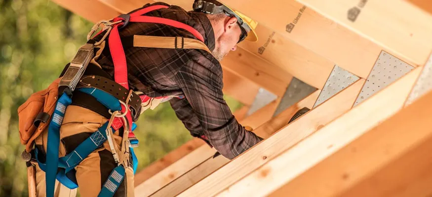 Construction worker wearing safety gear installs wooden beams on a roof structure outdoors.