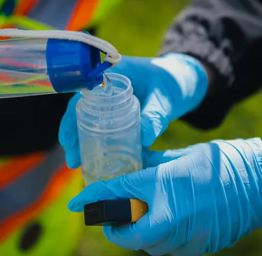Two gloved hands transfer liquid from a container into a smaller bottle outdoors.