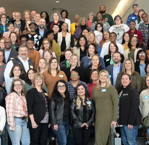 A large, diverse group of people stand and sit on a staircase inside a building, posing for a group photo. Some individuals wear nametags and are dressed casually.