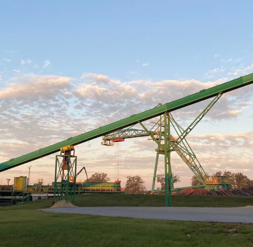 A green, industrial conveyor belt system is shown extending across an outdoor facility, with metal structures and large containers underneath a partly cloudy sky.