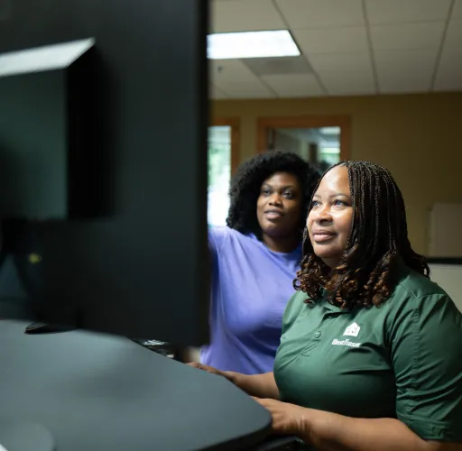 Two people are working at computers in an office. One is wearing a purple shirt and the other a green shirt. They appear focused on their tasks.