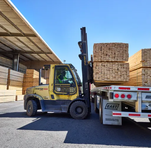 A forklift operator loads a large stack of lumber onto a flatbed trailer in an outdoor lumber yard, surrounded by stacks of wooden planks under a clear blue sky.