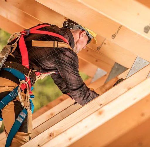 Construction worker wearing a safety harness, plaid shirt, and helmet, working on the wooden structure of a building.