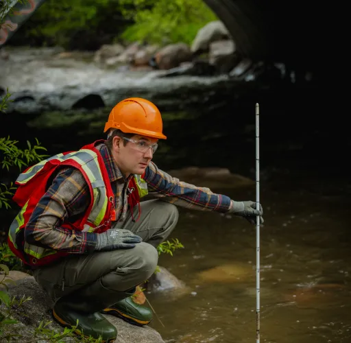 Person in a safety vest and helmet crouches by a stream, holding a measuring stick near a bridge.
