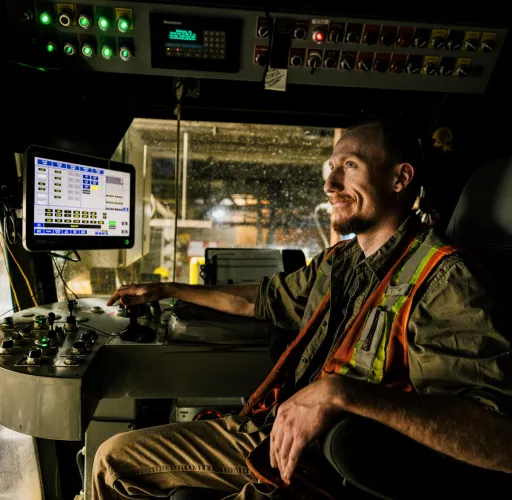 Person in a high-visibility vest operates machinery, sitting in a control room with multiple screens and control panels.