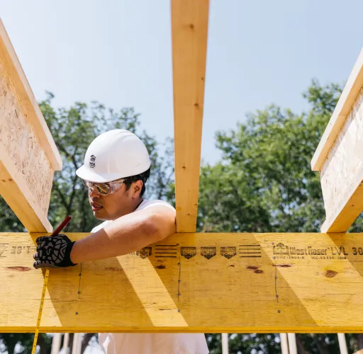A construction worker wearing a white hard hat and safety glasses measures and marks a wooden beam at a construction site.