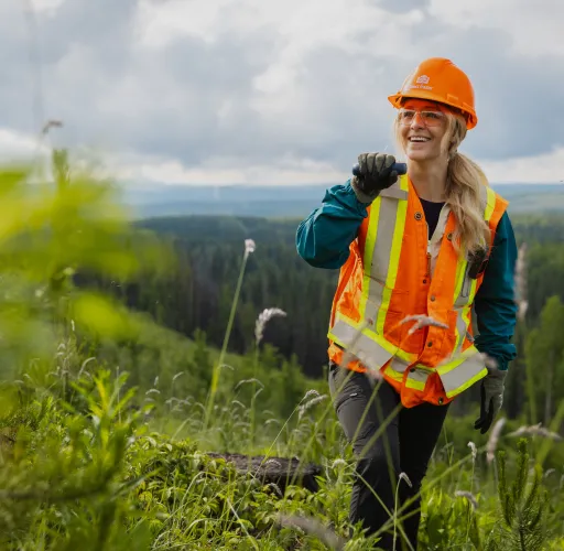 A person wearing an orange hard hat and safety vest stands in a green forested area, smiling and holding a radio.