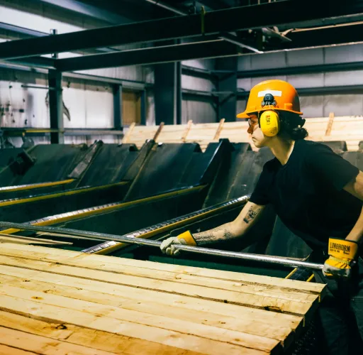 A worker wearing protective gear handles wooden planks in an industrial setting, with machinery visible in the background.