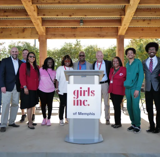 A group of people stands together under a wooden pavilion, with a podium labeled "Girls Inc. of Memphis" in front.