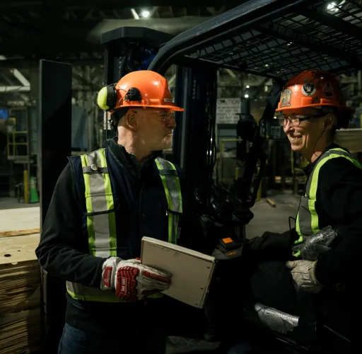 Two workers wearing hard hats and safety vests converse near a forklift in a lumber warehouse. One holds a tablet, and the other is seated on the forklift.