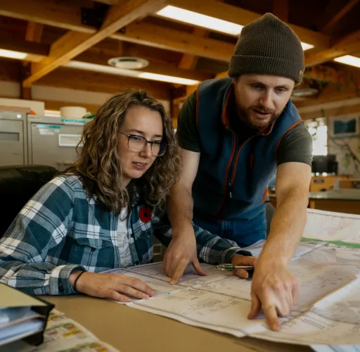 Two people reviewing large maps on a table. The woman is seated, wearing glasses, and the man is pointing at the map, wearing a beanie. They are in a room with wooden beams and filing cabinets.