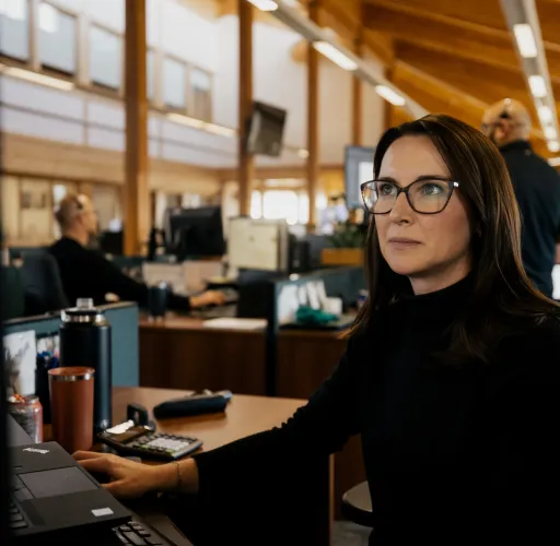 A woman with glasses works at a computer in an office with wooden ceilings. Other people are seen working in the background.