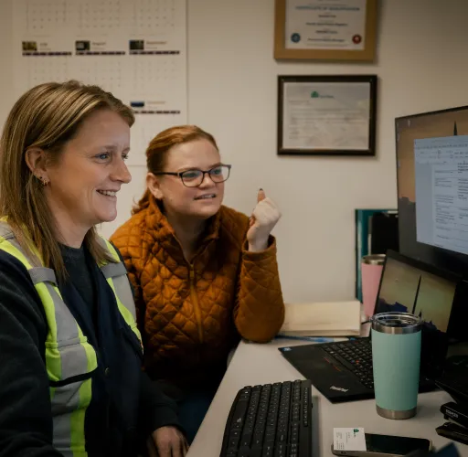 Two women sitting at a desk, looking at a computer screen. One wears a reflective vest. The wall behind them has a calendar and framed certificates.