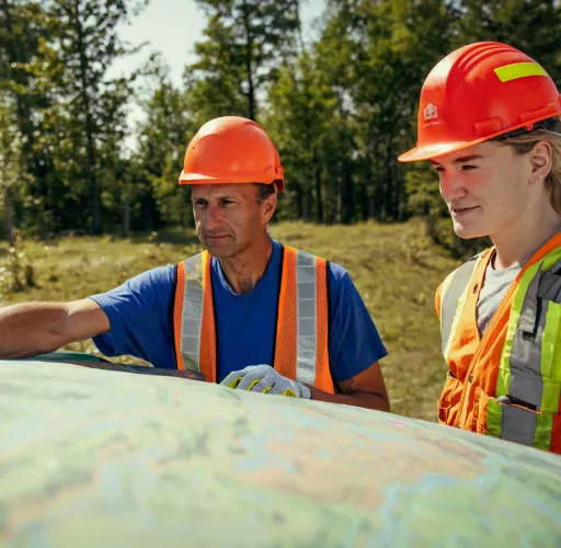 Two people wearing orange safety vests and hard hats look at a large map outdoors in a wooded area on a sunny day.