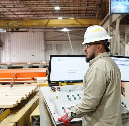 A worker wearing a hard hat and gloves operates a control panel with screens and buttons inside an industrial facility.