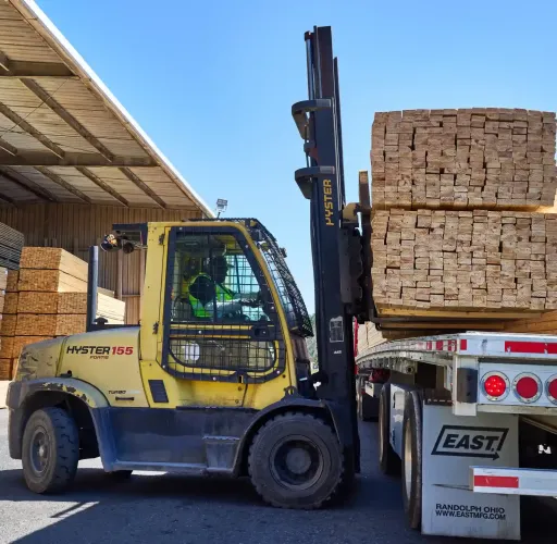 A forklift operator loads stacked lumber onto a flatbed trailer in an outdoor lumber yard under clear skies.